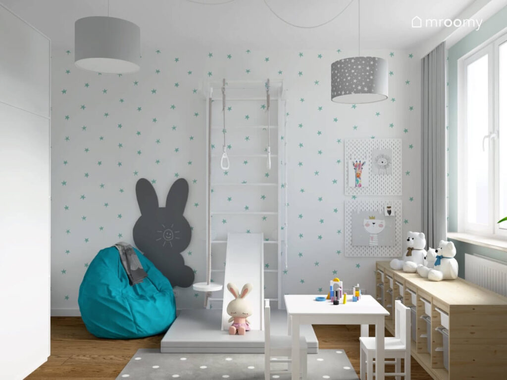 Biała ściana pokryta tapetą w niebieskie gwiazdki a także niebieski worek do siedzenia drabinka gimnastyczna ze zjeżdżalnią i szara tablica kredowa w kształcie królika w pokoju dla dziewczynki
