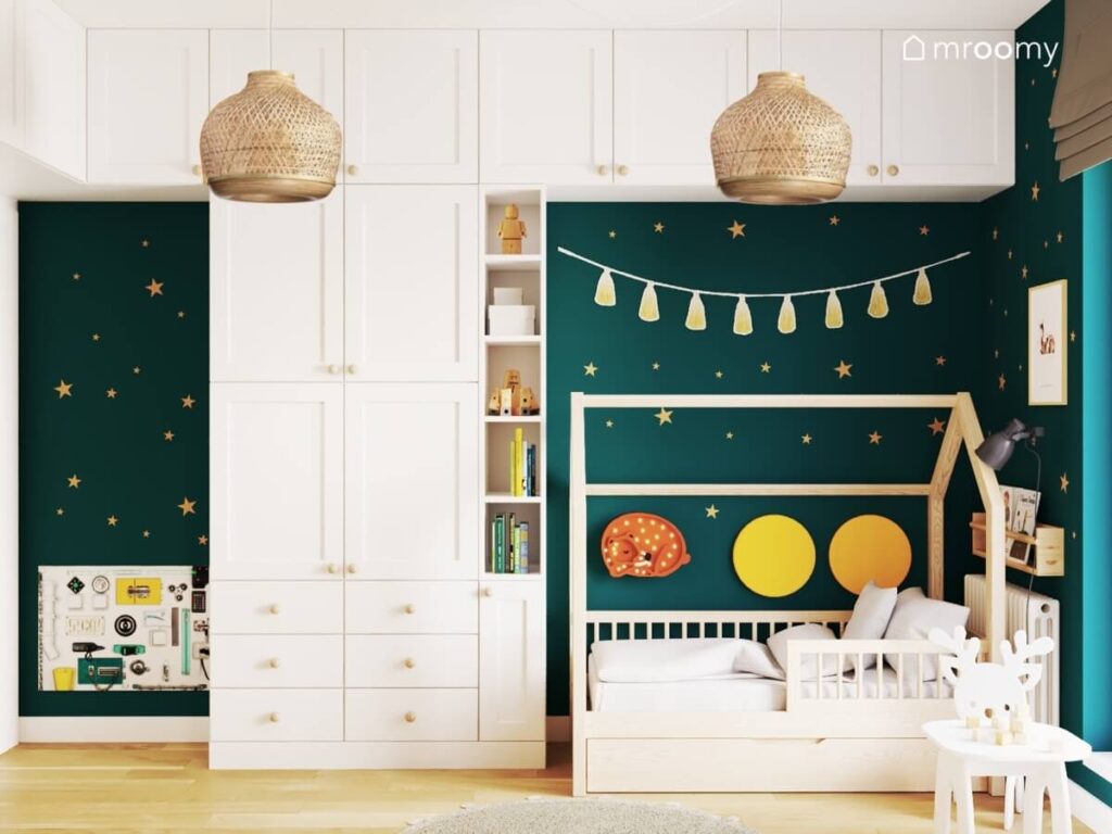 Białe meble modułowe z drewnianymi gałkami oraz drewniane łóżko domek uzupełnione miękkimi panelami i lampką nocną w kształcie sarenki a za nimi zielona ściana w gwiazdki w pokoju dla dwóch braci
