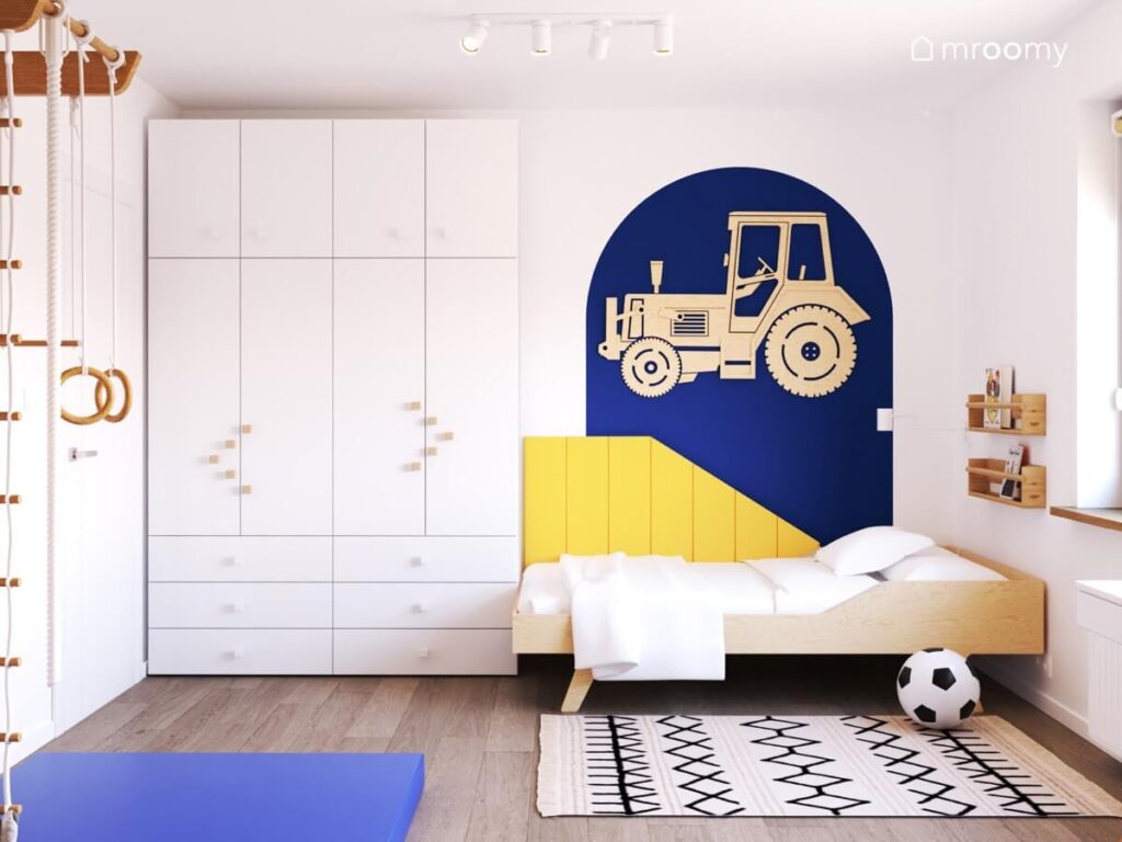 Biały pokój dla chłopca a w nim biała szafa drewniane łóżko a za nim żółte panele ścienne i traktor ze sklejki