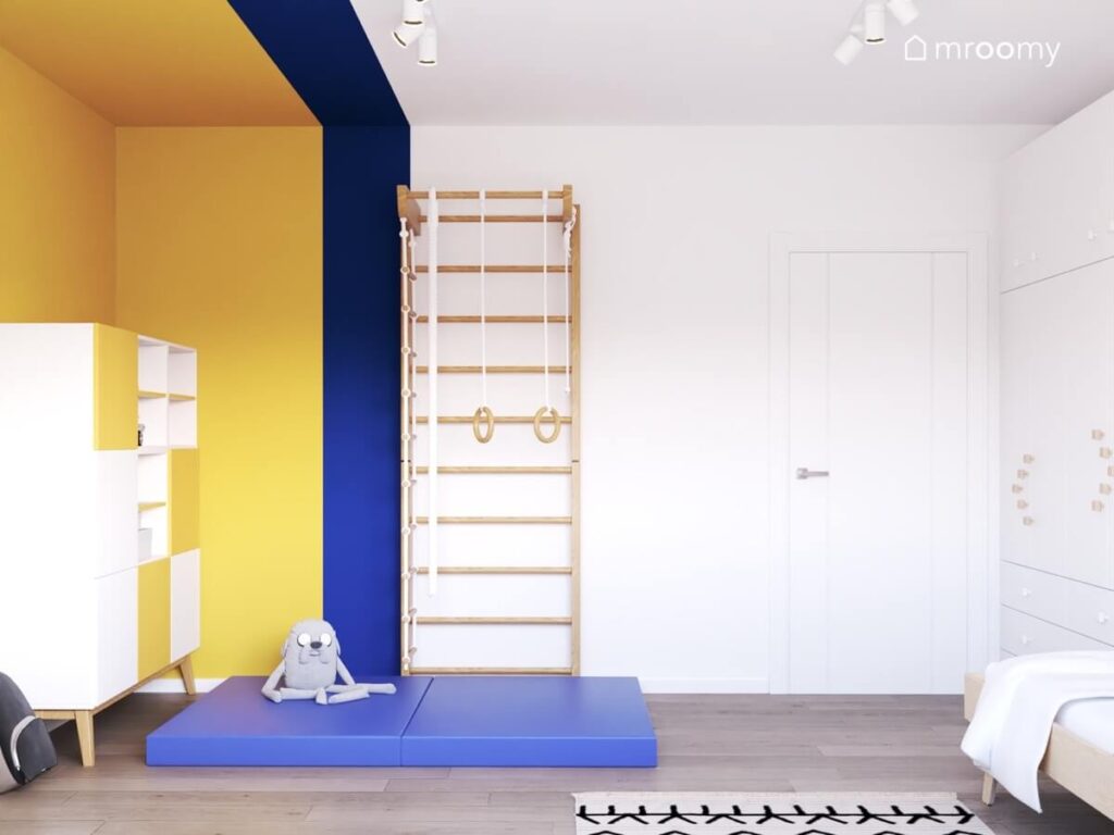 Drewniana drabinka gimnastyczna z niebieskimi materacami oraz biało żółty regał w biało żółto niebieskim pokoju dla chłopca w wieku szkolnym