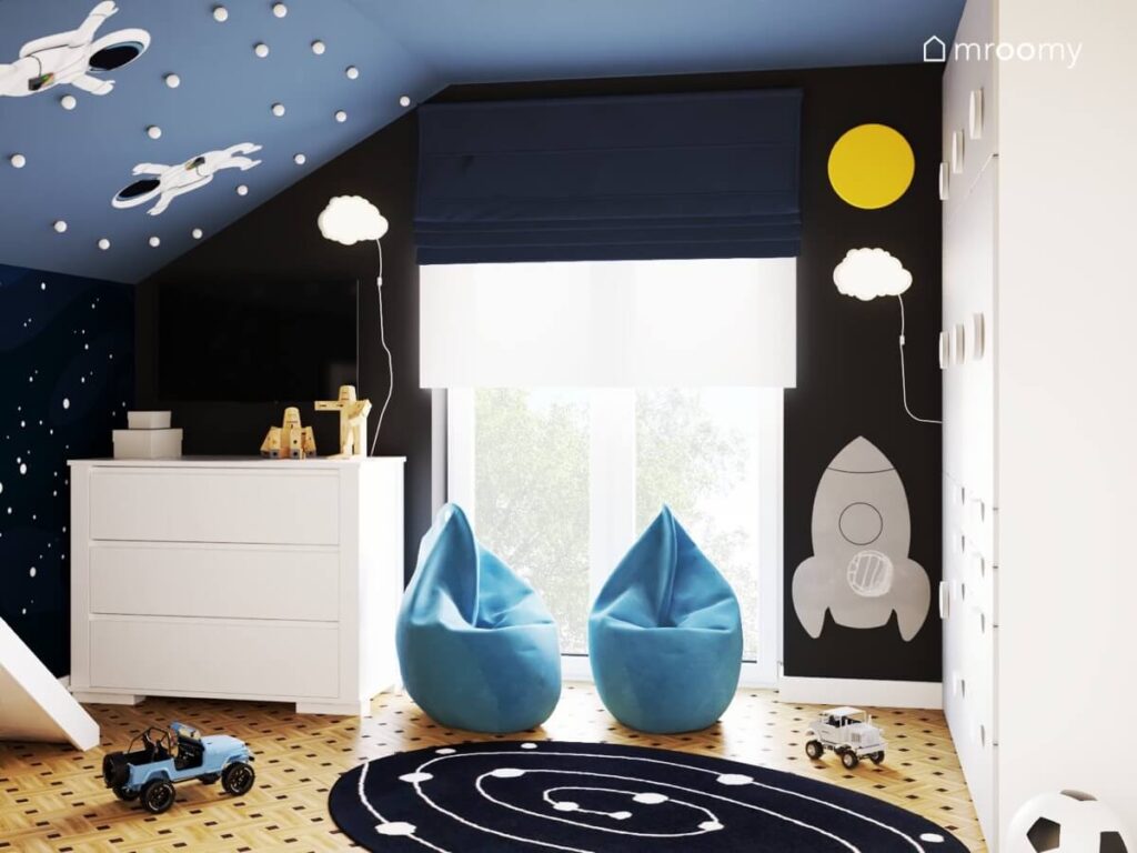 Dwie niebieskie pufy tablica kredowa w kształcie lakiery lampki ścienne w kształcie chmurek oraz biała komoda w czarno niebieskim pokoju dla chłopców