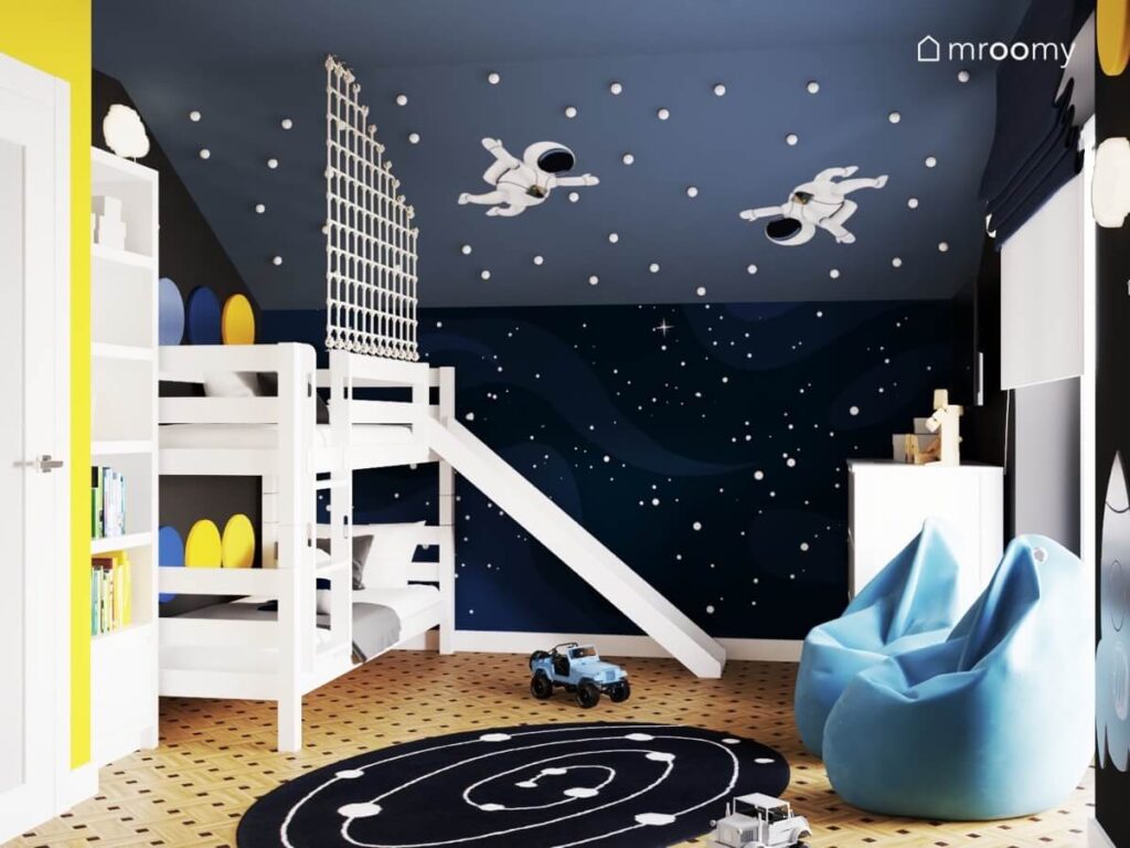 Kosmiczny pokój dla dwóch chłopców z białym łóżkiem piętrowym dwoma pufami dywanem z układem planetarnym oraz ze ścianą ozdobioną naklejkami w kształcie kosmonautów oraz gałkami imitującymi gwiazdy