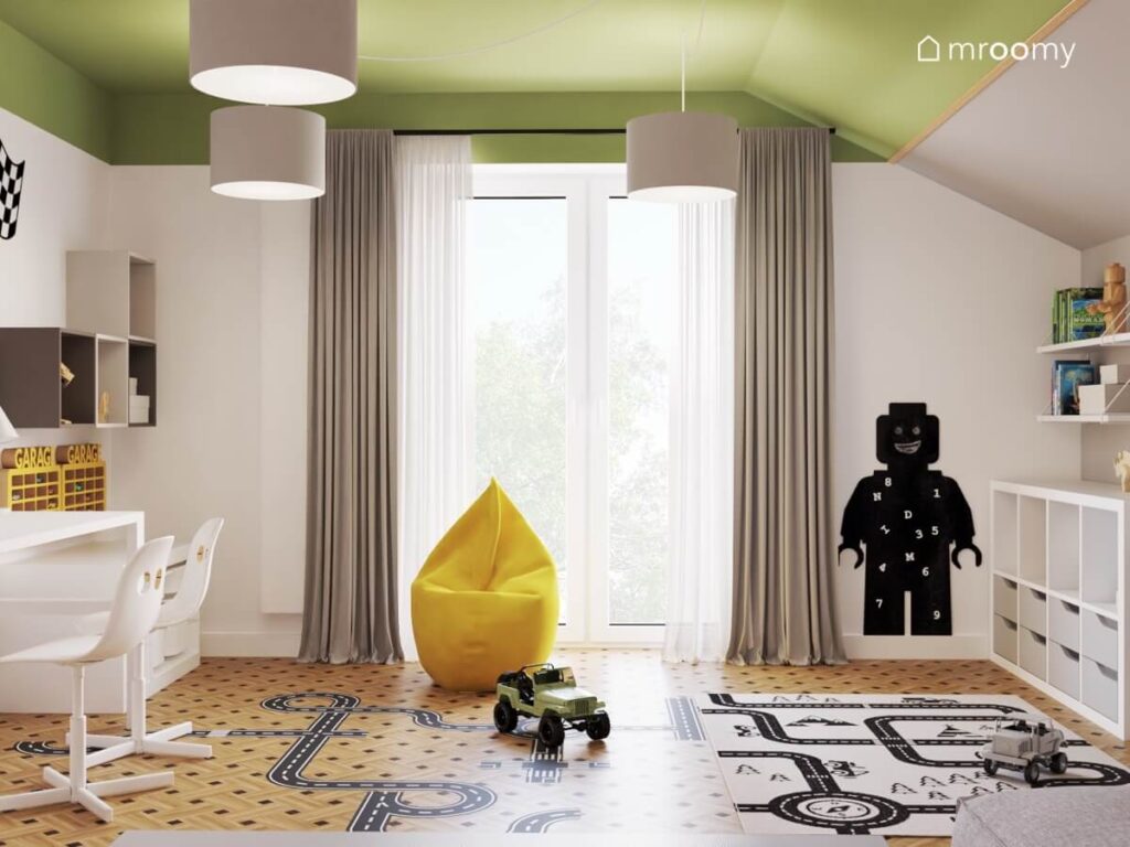 Szaro zielony pokój zabaw dla dwóch chłopców z tablicą kredową w kształcie ludzika Lego żółtą pufą oraz dywanem z motywem miasta i naklejką podłogową w kształcie jezdni