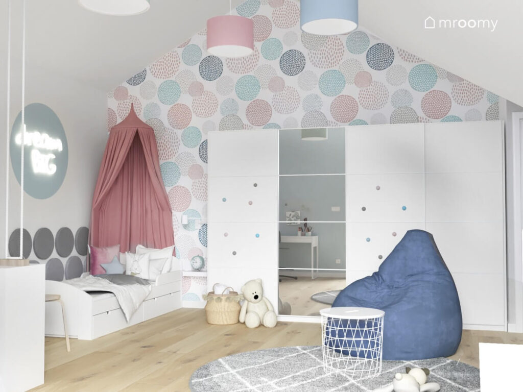 Ściana pokryta kolorową tapetą a także biała szafa z lustrzanym fontem uzupełniona kolorowymi gałkami oraz niebieska pufa i łóżko uzupełnione różowym baldachimem w pokoju dziewczynki