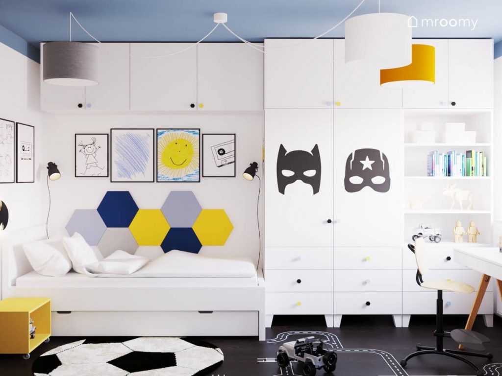 Biało niebieski pokój dla chłopca a w nim szafa uzupełniona naklejkami w kształcie masek superbohaterów łóżko z kolorowymi panelami ściennymi oraz galeria dziecięcych rysunków w ramkach a na suficie lampa z kolorowymi abażurami