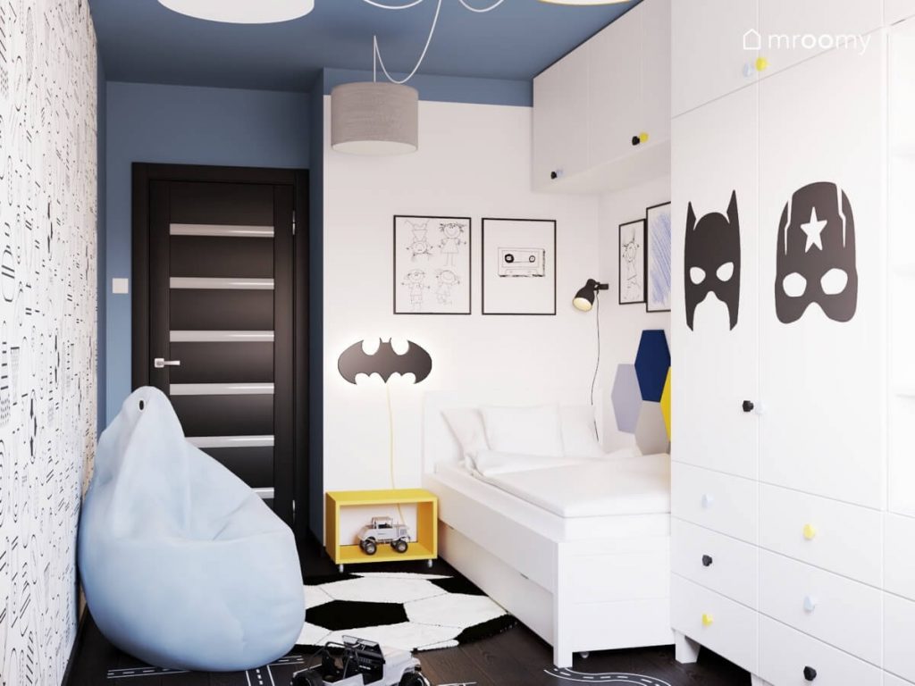 Biały pokój dla chłopca z niebieskim sufitem dywanem w kształcie piłki i białym łóżkiem z lampką nocną w kształcie logo Batmana i żółtym stolikiem nocnym na kółkach