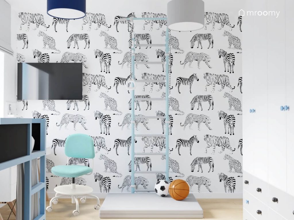Ściana pokryta tapetą w zebry i dzikie koty a na niej błękitna drabinka gimnastyczna z szarym materacem w pokoju dla chłopca