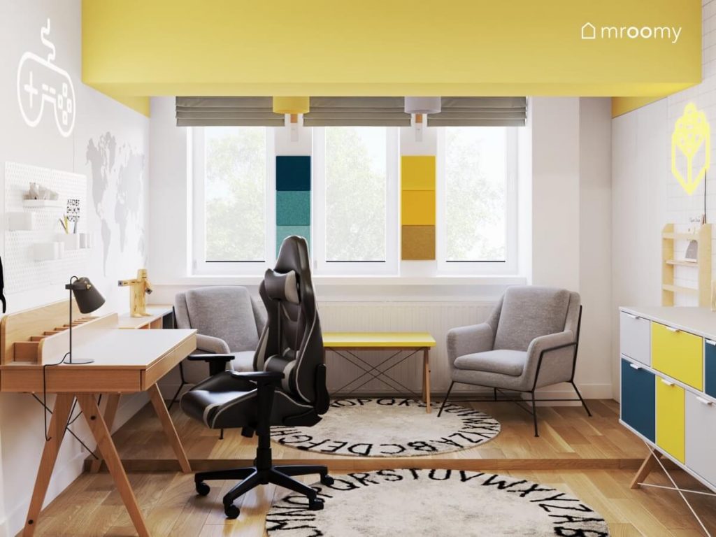 Drewniane biurko z fotelem gamingowym oraz żółty stolik z szarymi fotelami a na podłodze dywany z alfabetem w biało żółtym pokoju chłopca