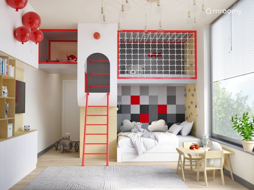 Pokój dla kilkulatka z rozbudowaną antresolą na którą prowadzi czerwona drabina oraz ścianka wspinaczkowa a pod nią znajduje się łóżko uzupełnione szarymi i czerwonymi panelami ściennymi