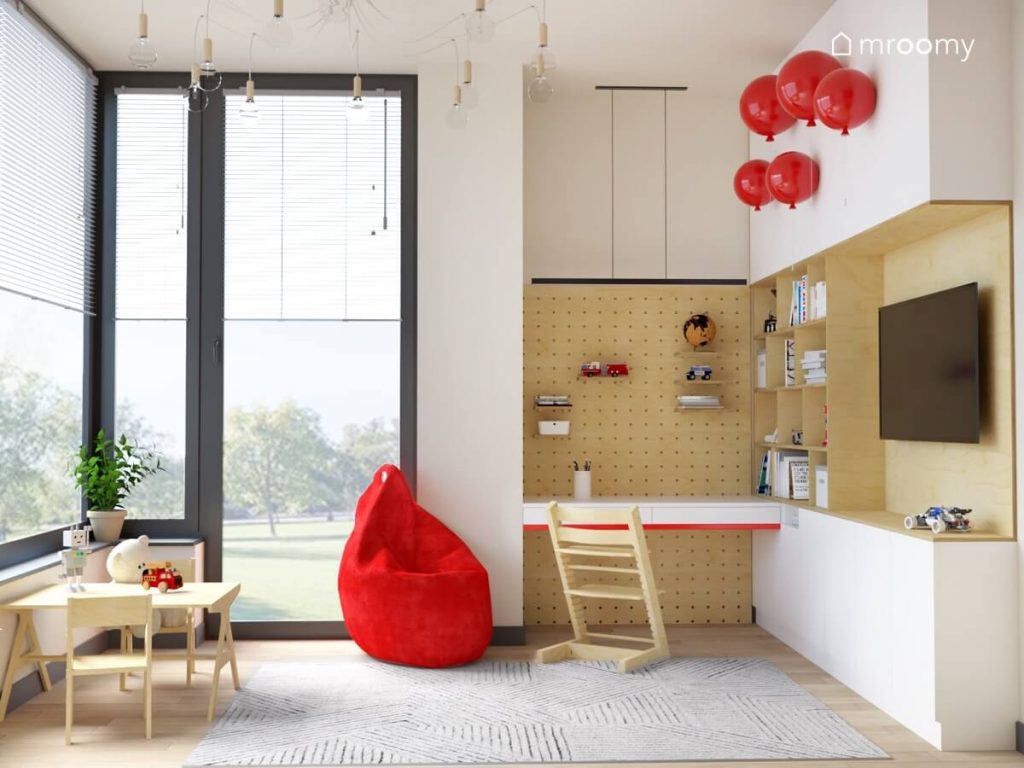 Jasny pokój dla chłopca z biało drewnianą zabudową meblową niskim stolikiem z krzesełkiem czerwoną pufą sako oraz z oryginalnym oświetleniem w postaci lamp balonów oraz żyrandola z licznymi żarówkami