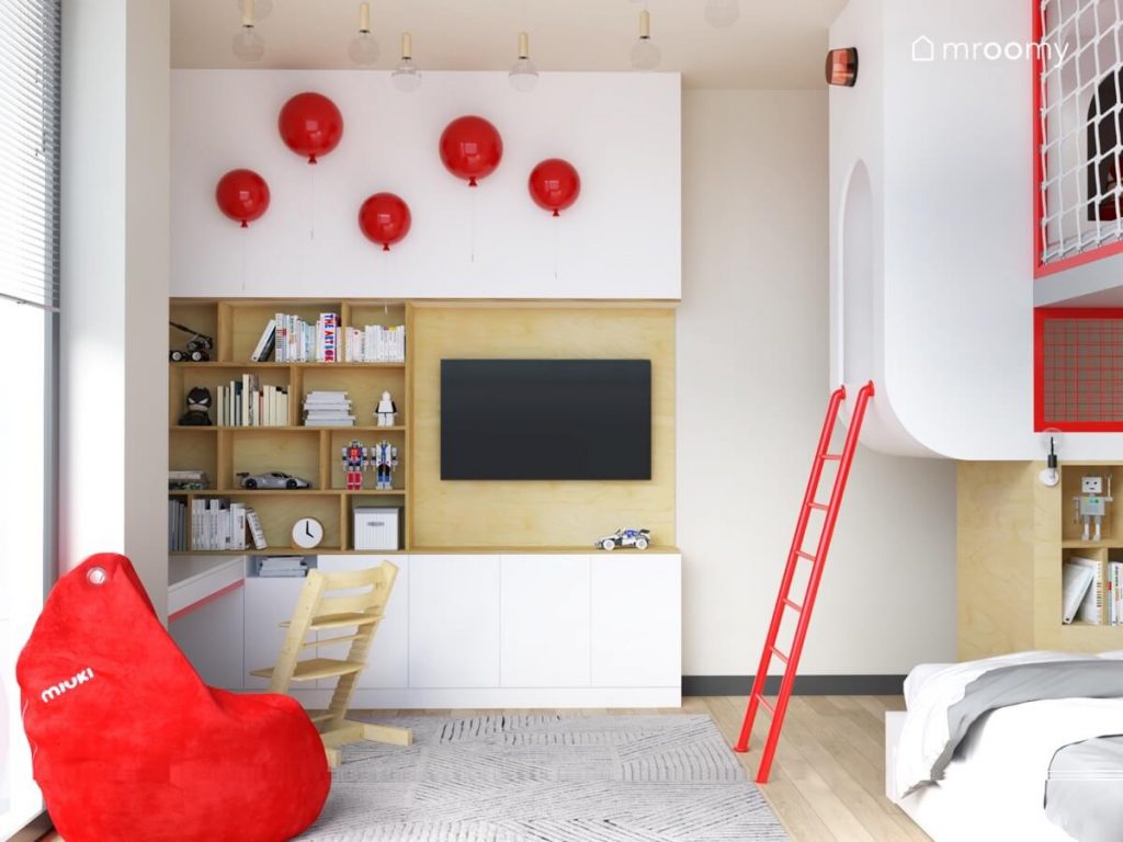 Biało drewniana zabudowa meblowa w pokoju dla chłopca ozdobiona kinkietami w kształcie balonów a także czerwona pufa sako i antresola na którą prowadzi czerwona drabina