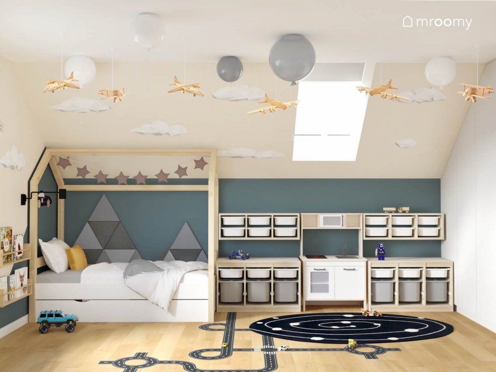 Niebiesko biały poddaszowy pokój dla chłopca a w nim drewniane łóżko domek uzupełnione gwieździstą girlandą i panelami ściennymi a obok kuchenka dla dzieci i drewniane regały z białymi i szarymi pojemnikami