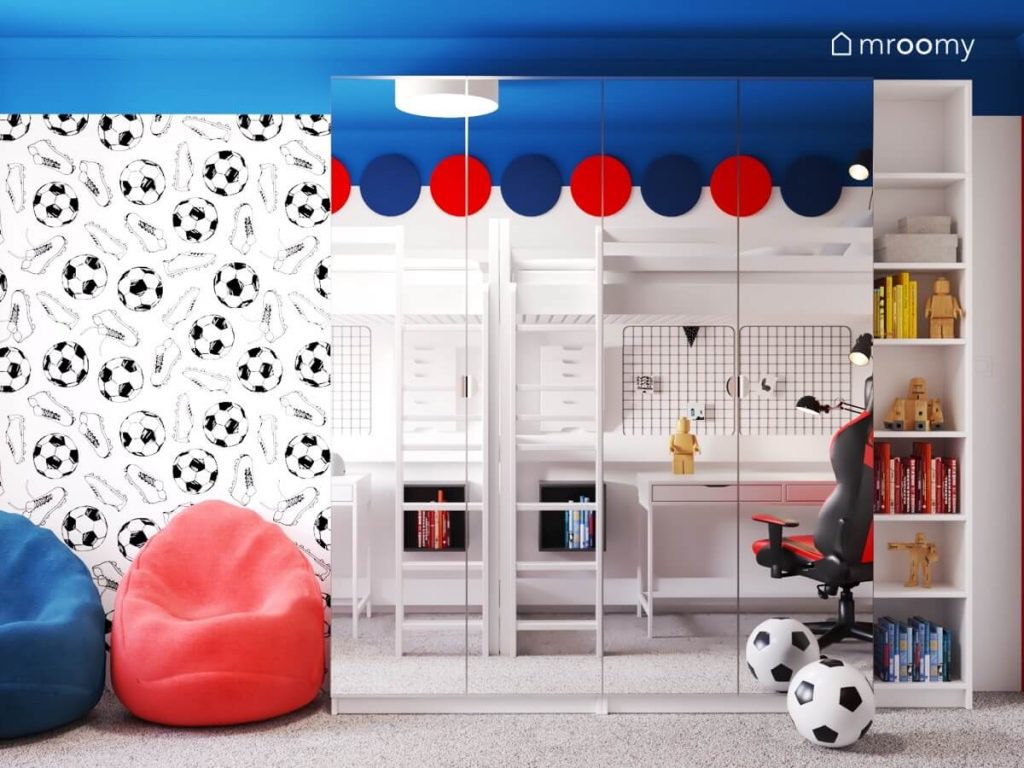 Szafa z lustrzanymi frontami a także niebieski i czerwony worek do siedzenia a na ścianie piłkarska tapeta w pokoju dla dwóch chłopców w wieku szkolnym