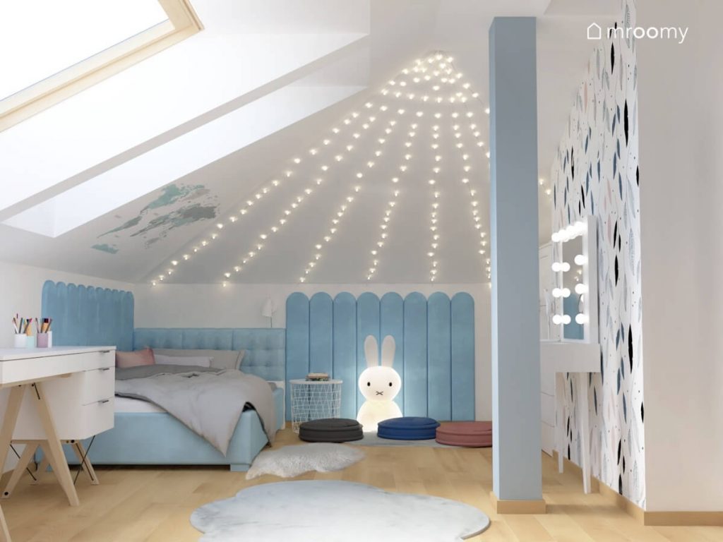 Niebieskie tapicerowane łóżko uzupełnione panelami ściennymi a na skosie kurtyna świetlna a na podłodze lampa w kształcie królika trzy dywany i trzy pufy francuskie w pokoju dziewczynki