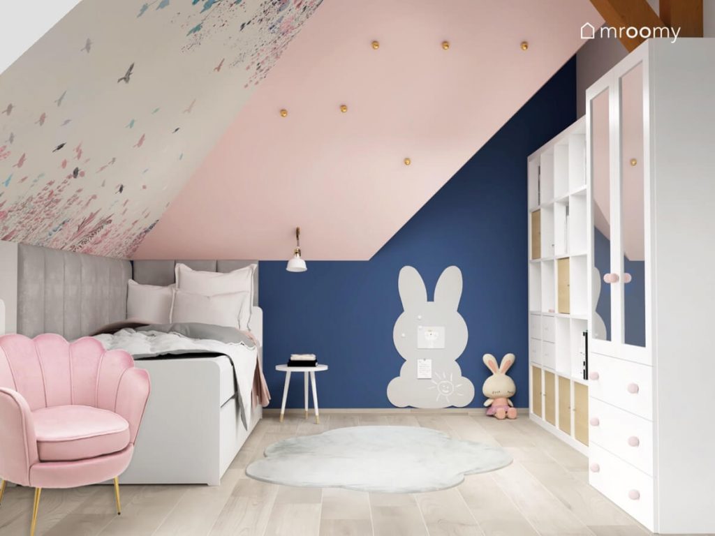 Łóżko uzupełnione szarymi panelami ściennymi tablica kredowa w kształcie królika oraz szary dywanik w kształcie chmurki w różowo granatowym pokoju dziewczynki