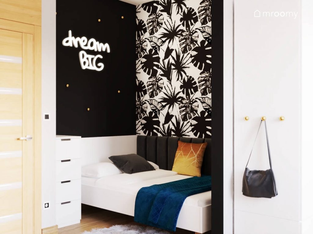 Strefa spania w czarno biało szarym pokoju dla nastolatki a w niej łóżko uzupełnione czarnymi panelami ściennymi tapeta w egzotyczne liście oraz ledon w kształcie napisu