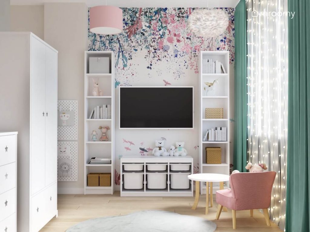Ściana multimedialna w pokoju dla rocznej dziewczynki a na niej telewizor dwa wysokie regały regał z pojemnikami na zabawki oraz kwiecista tapeta