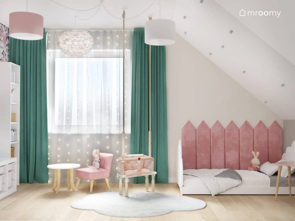 Jasny poddaszowy pokój dla małej dziewczynki a w nim białe łóżko uzupełnione różowymi panelami ściennymi a także huśtawka wisząca stolik z fotelikiem oraz okno ozdobione miętowymi zasłonami i kurtyną świetlną