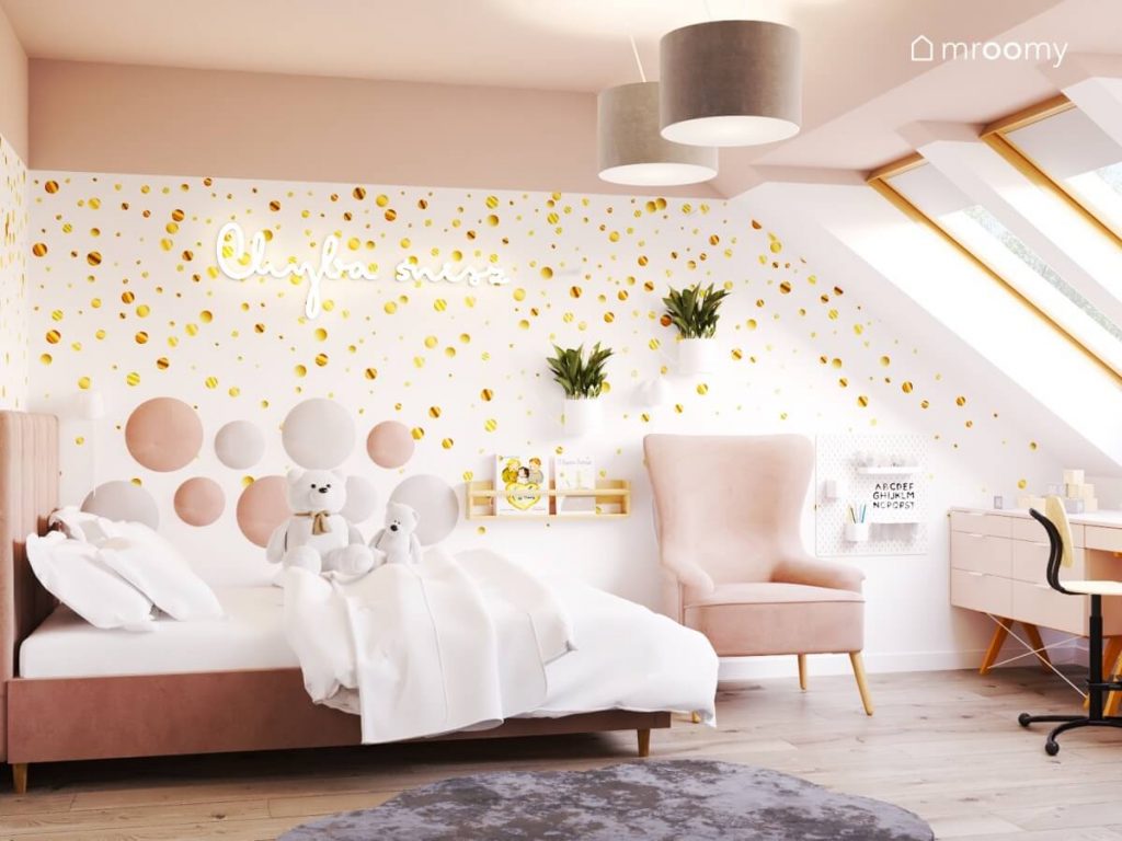 Strefa spania w pokoju kilkuletniej dziewczynki a w niej różowe welurowe łóżko oraz ściana ozdobiona tapetą w złote kropki miękkimi panelami ledonem w kształcie napisu oraz kwietnikami