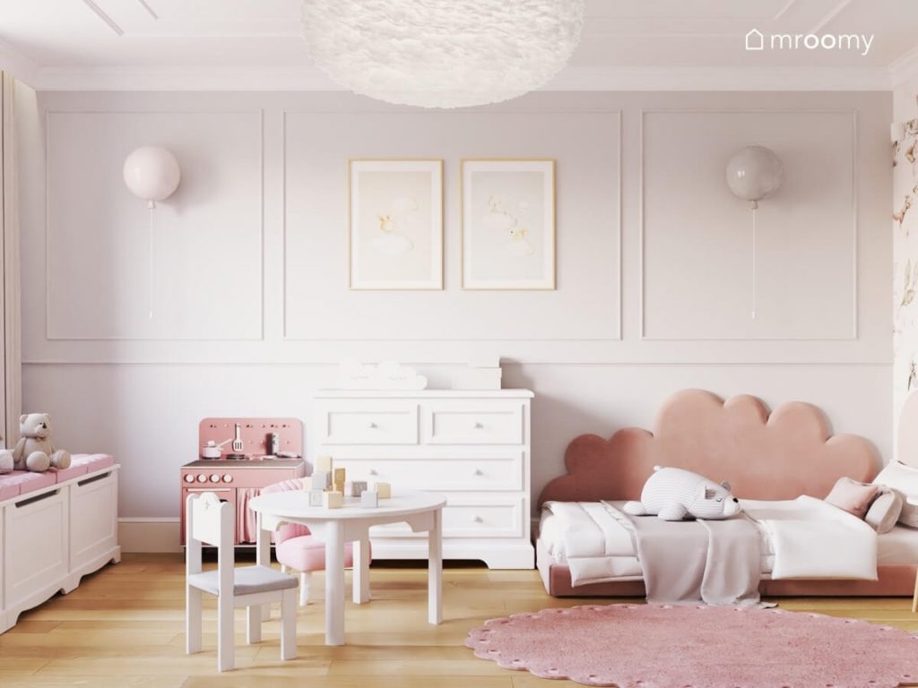 Szara ściana w pokoju dziewczynki noworodka a na niej ozdobna sztukateria kinkiety w kształcie balonów oraz plakaty w ramkach a pod spodem łóżko z oparciem w kształcie chmurki komoda oraz kuchenka dla dzieci