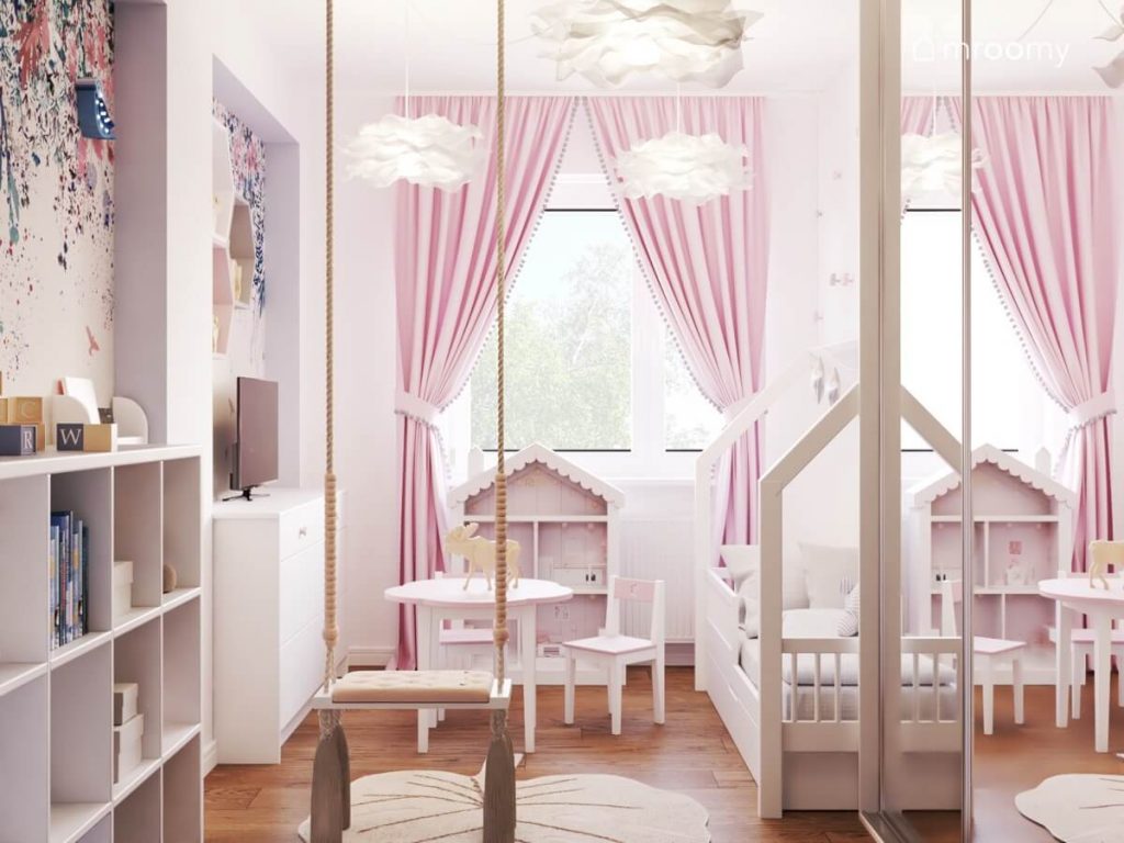 Jasny pokój dla małej dziewczynki a w nim huśtawka z frędzlami łóżko domek oraz regał w kształcie domku a także różowe zasłony i ozdobne materiałowe lampy