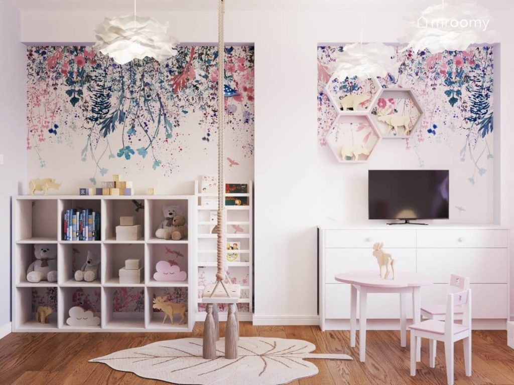 Ściana pokryta kwiecistą tapetą w pokoju małej dziewczynki a na niej telewizor półka w kształcie heksagonów biblioteczka oraz kwadratowy regał