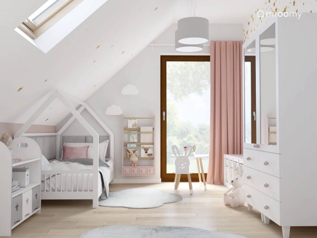Jasny poddaszowy pokój dla dziewczynki a w nim łóżko domek różowa biblioteczka lampki ścienne i dywaniki w kształcie chmurek oraz stolik z krzesełkiem w kształcie królika