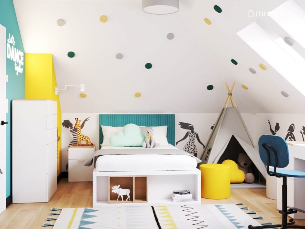 Strefa spania w pokoju dla kilkulatki z łóżkiem uzupełnionym niebieskimi panelami ściennymi i dębowymi skrzyniami a także z namiotem tipi lampką nocną w kształcie żyrafy i skosem z kolorowymi wieszakami