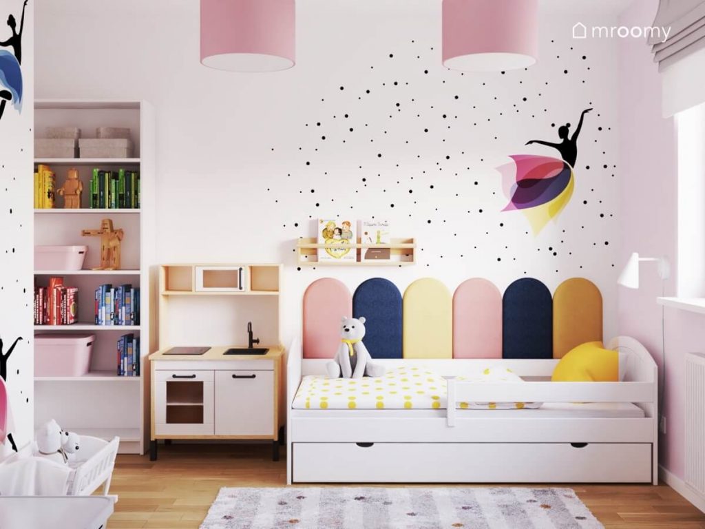 Białe łóżko uzupełnione panelami ściennymi w różnych kolorach a także kuchnia dla dzieci i biały regał a na ścianie naklejki w kształcie baletnicy i kropek w pokoju dziewczynki