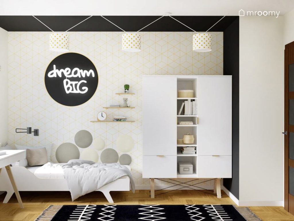 Ściana pokryta jasną geometryczną tapetą i miękkimi panelami a także białe łóżko ledon w kształcie napisu i szafa na drewnianych nogach w pokoju nastolatki