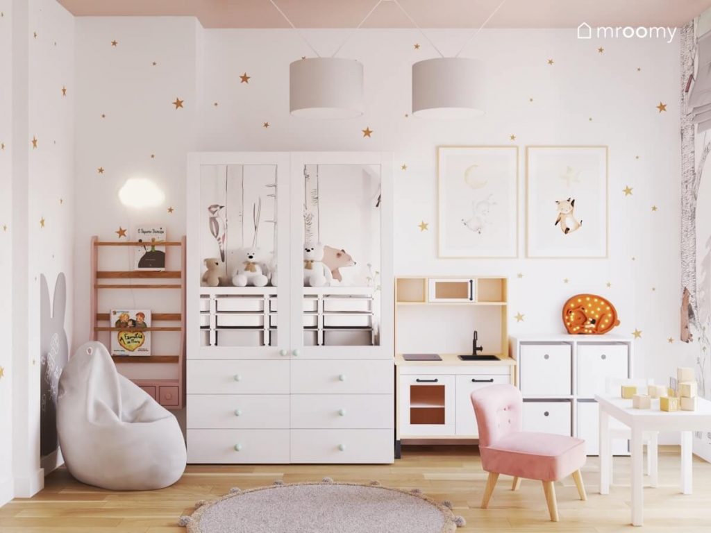 Różowa biblioteczka biała szafa z lustrzanymi frontami kuchnia dla dzieci a na ścianie tablica kredowa w kształcie królika ozdobne plakaty lampka w kształcie chmurki oraz złote gwiazdki w pokoju malutkiej dziewczynki