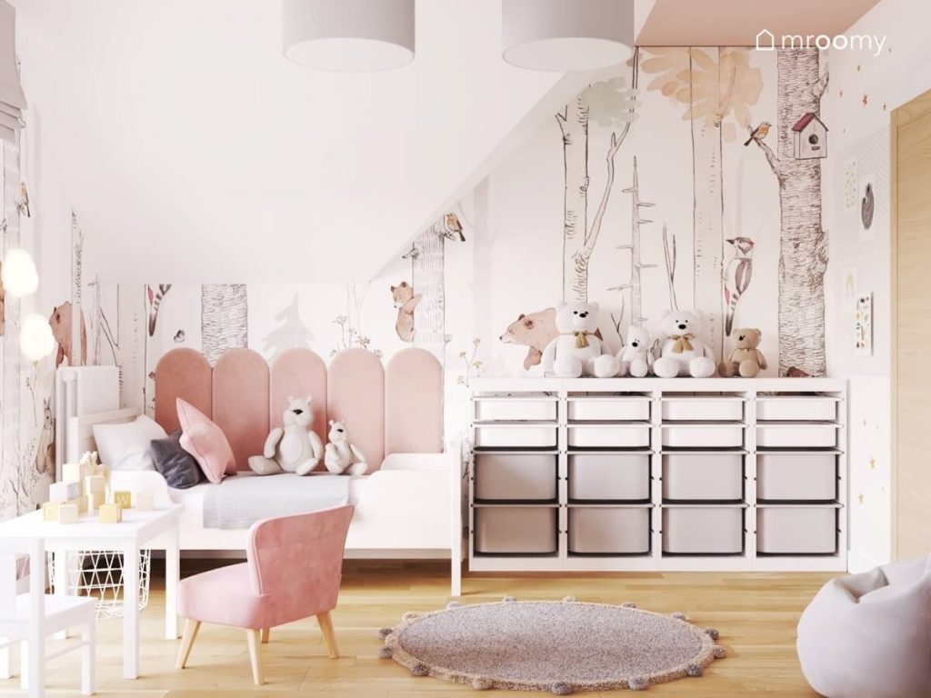 Białe łóżko uzupełnione różowymi panelami ściennymi a obok regał z białymi i szarymi pojemnikami a na ścianie leśna tapeta w pokoju dla dziewczynki