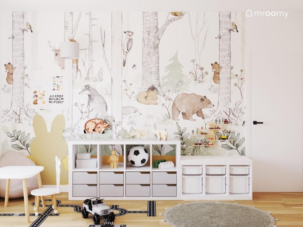 Ściana pokryta tapetą z leśnym motywem oraz regały z pojemnikami i tablica kredowa w kształcie królika w pokoju siostry i brata