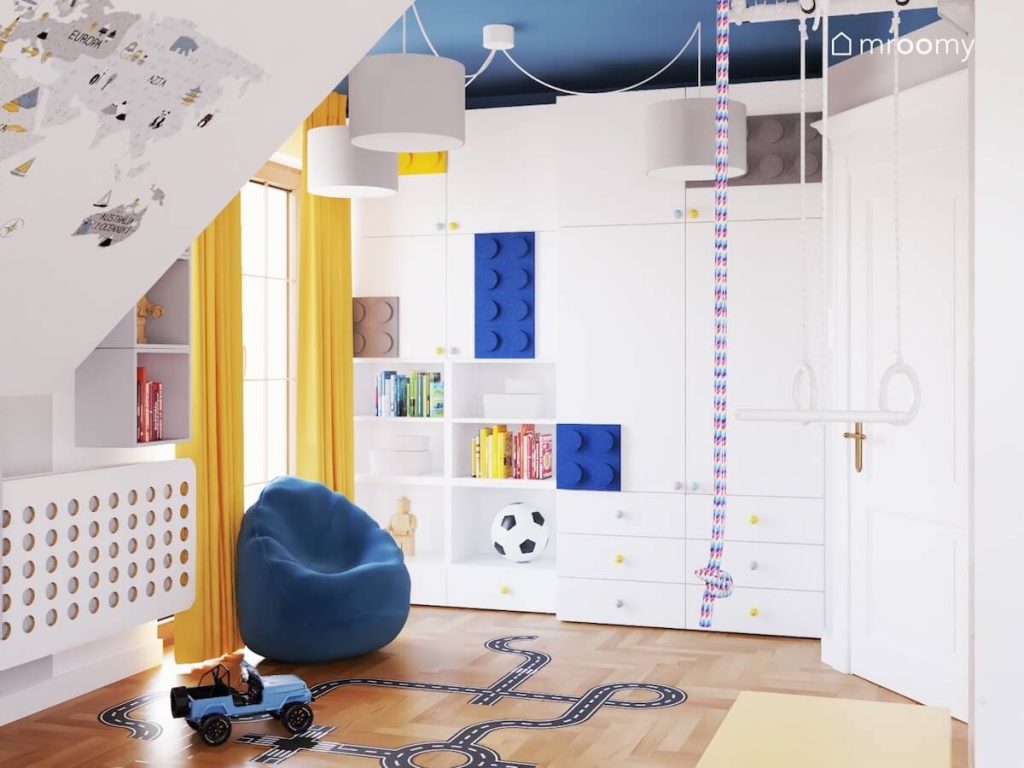 Biała szafa z kolorowymi gałkami oraz panelami w kształcie klocków a także żółte zasłony niebieska pufa oraz biała lampa wisząca w pokoju dla chłopca