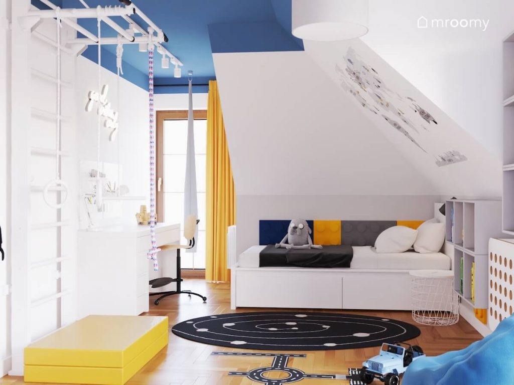 Biało niebieski poddaszowy pokój dla chłopca w wieku szkolnym a w nim białe meble drabinka gimnastyczna łóżko uzupełnione panelami w kształcie klocków a na podłodze naklejka w kształcie ulicy oraz kosmiczny dywan