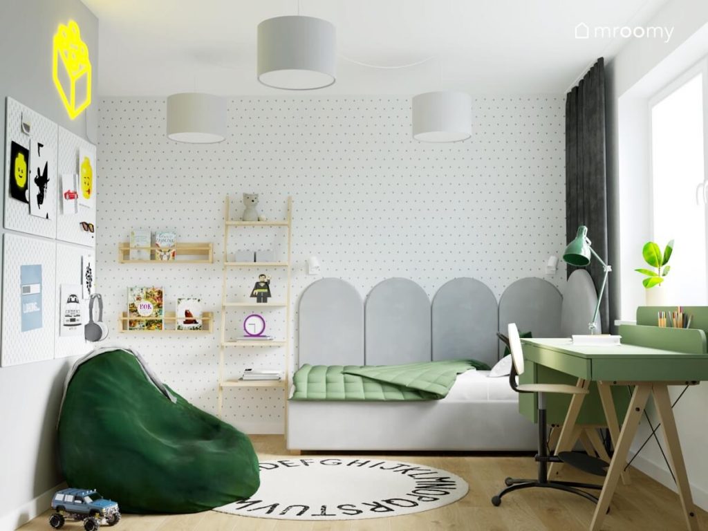 Pokój chłopca w wieku szkolnym utrzymany w tonacji biało szaro zielonej a w nim szare tapicerowane łóżko drewniane półki ścienne lampa sufitowa na rozłożystym zawieszeniu oraz tapeta w kropki