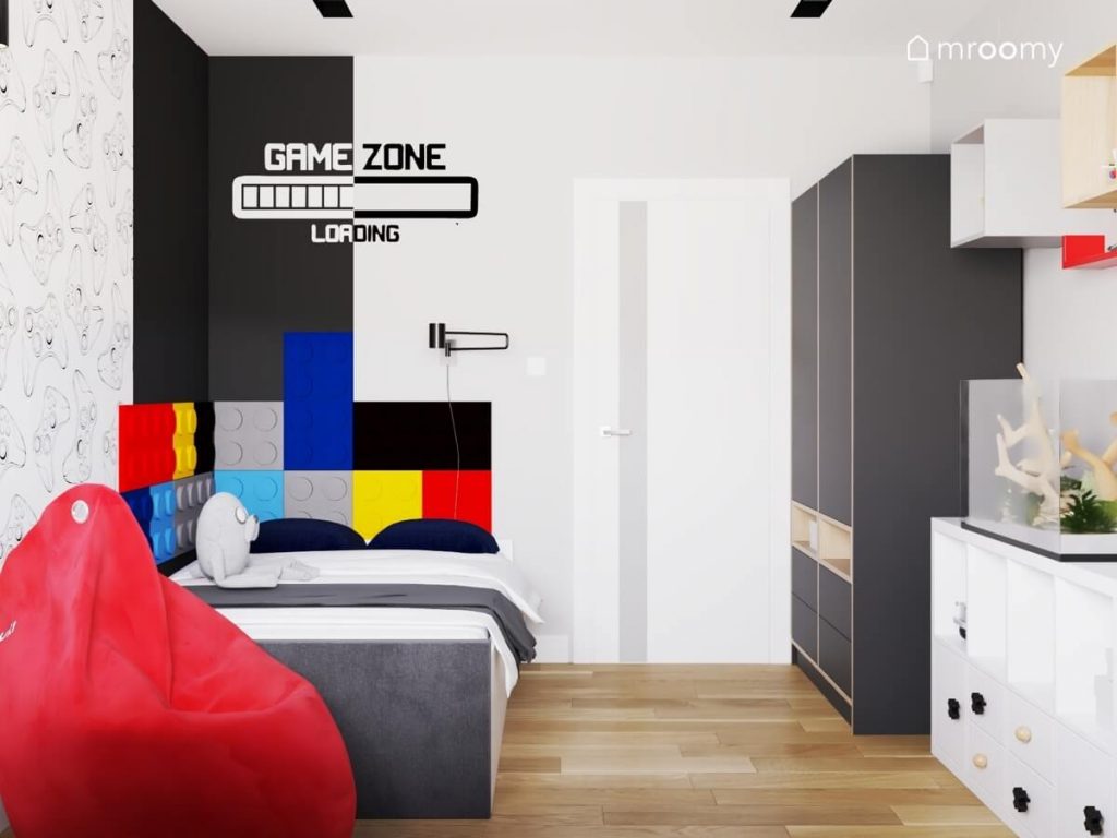 Biało czarny pokój dla chłopca w wieku szkolnym a w nim szara szafa łóżko uzupełnione kolorowymi panelami w kształcie klocków a na ścianie gamingowe motywy
