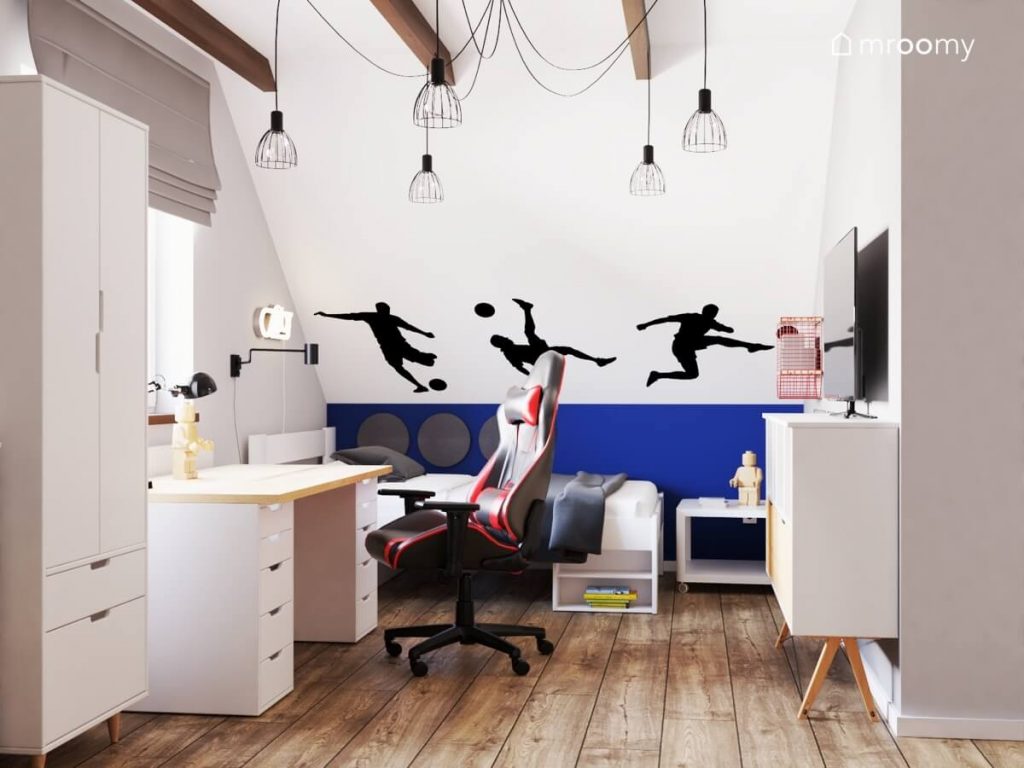 Strefa nauki w poddaszowym pokoju dla nastolatka a w niej biało drewniane biurko i fotel gamingowy a na ścianie naklejki w kształcie piłkarzy