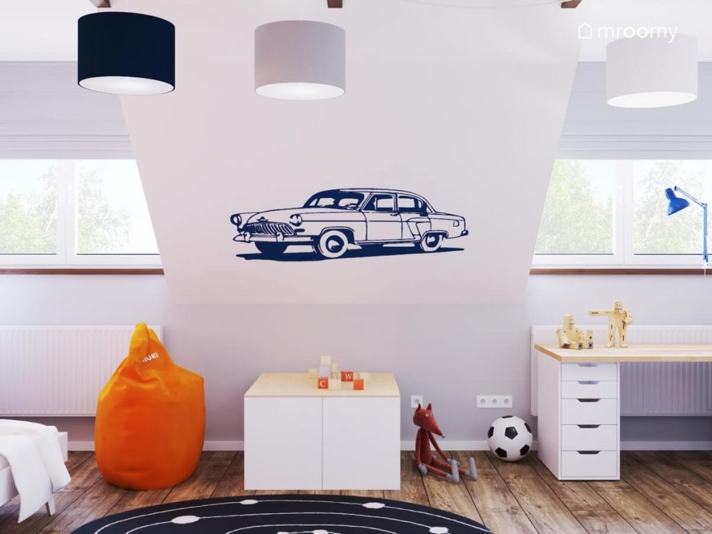 Skos ozdobiony naklejką w kształcie samochodowego klasyka a także biało drewniane meble pomarańczowa pufa oraz lampy sufitowe w trzech różnych abażurach w pokoju dla chłopca