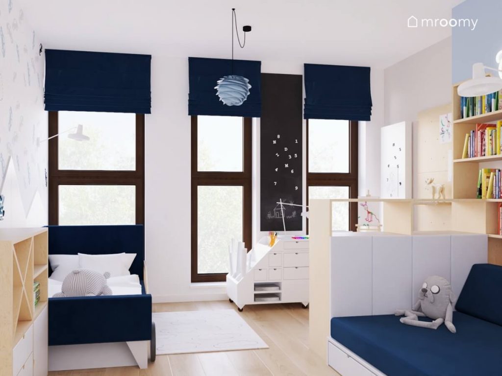 Biało szaro niebieski pokój dla chłopca w wieku przedszkolnym a w nim białe i drewniane meble oraz granatowe łóżko kanapa oraz rolety