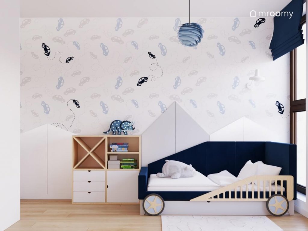 Strefa spania w pokoju dla czterolatka a w niej łóżko w kształcie samochodu szafka ze sklejki a na ścianie panele ścienne w kształcie gór lampka w kształcie triceratopsa i tapeta w autka