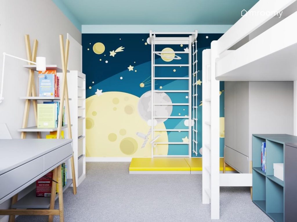 Szary pokój dla chłopca z błękitnym sufitem białą drabinką gimnastyczną z żółtym materacem białym łóżkiem na antresoli oraz kosmiczną tapetą na jednej ze ścian