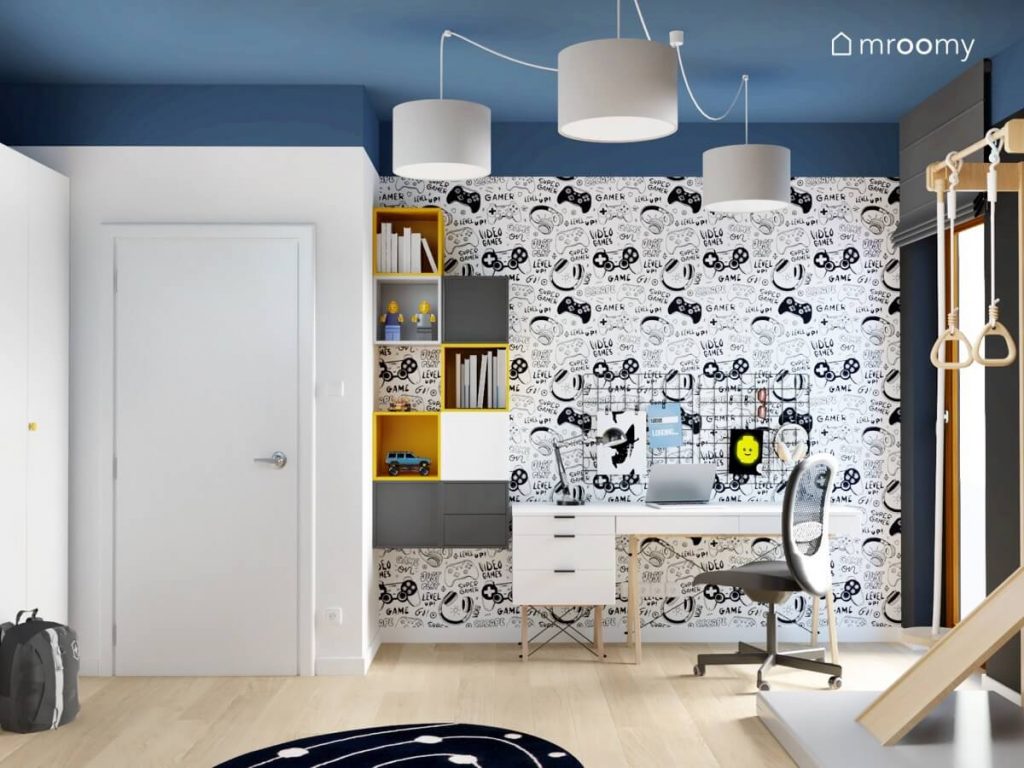 Strefa nauki w niebiesko biało czarnym pokoju dla chłopca a w nim białe biurko na drewnianych nogach z organizerami i szafkami ściennymi a na ścianie tapeta z motywem gamingowym