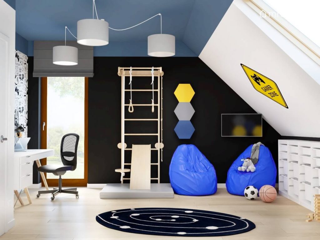 Czarno niebiesko biały poddaszowy pokój dla chłopca a w nim drabinka gimnastyczna ze zjeżdżalnią dwa niebieskie worki sako a także galaktyczny dywan i naklejka gamingowa na skosie