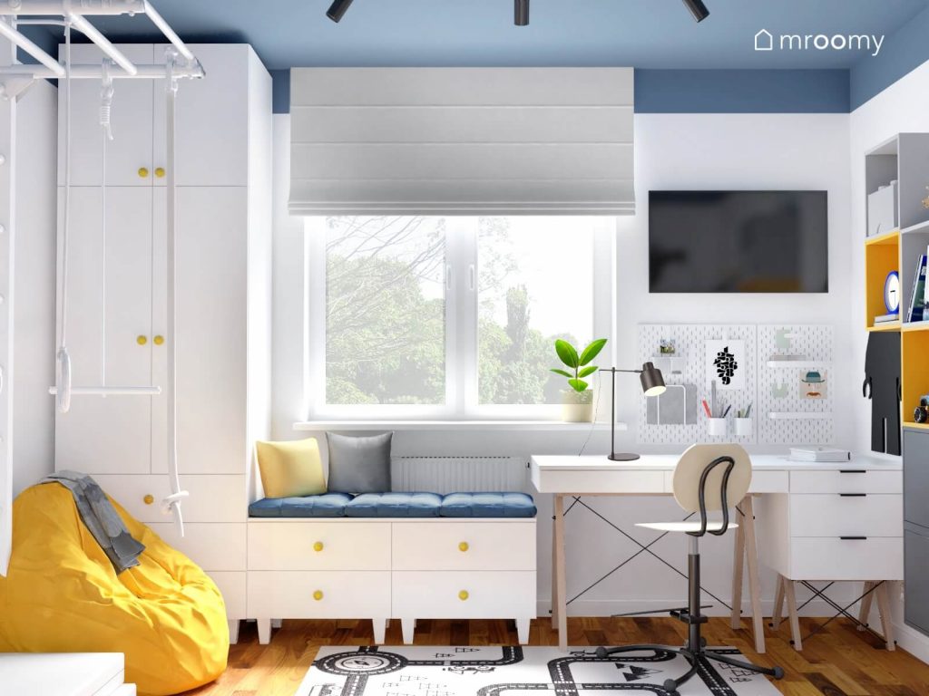 Białe meble modułowe z żółtymi gałkami a także białe biurko z kontenerkiem na drewnianych nogach a na ścianie organizery i telewizor w biało niebieskim pokoju chłopca