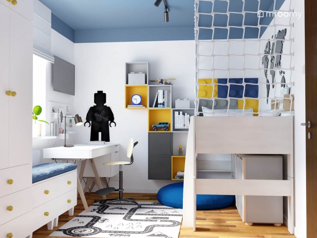 Biało niebieski pokój dla chłopca a w nim białe biurko na drewnianych nogach szare i żółte szafki ścienne dywan z motywem miasta oraz tablica kredowa w kształcie ludzika Lego
