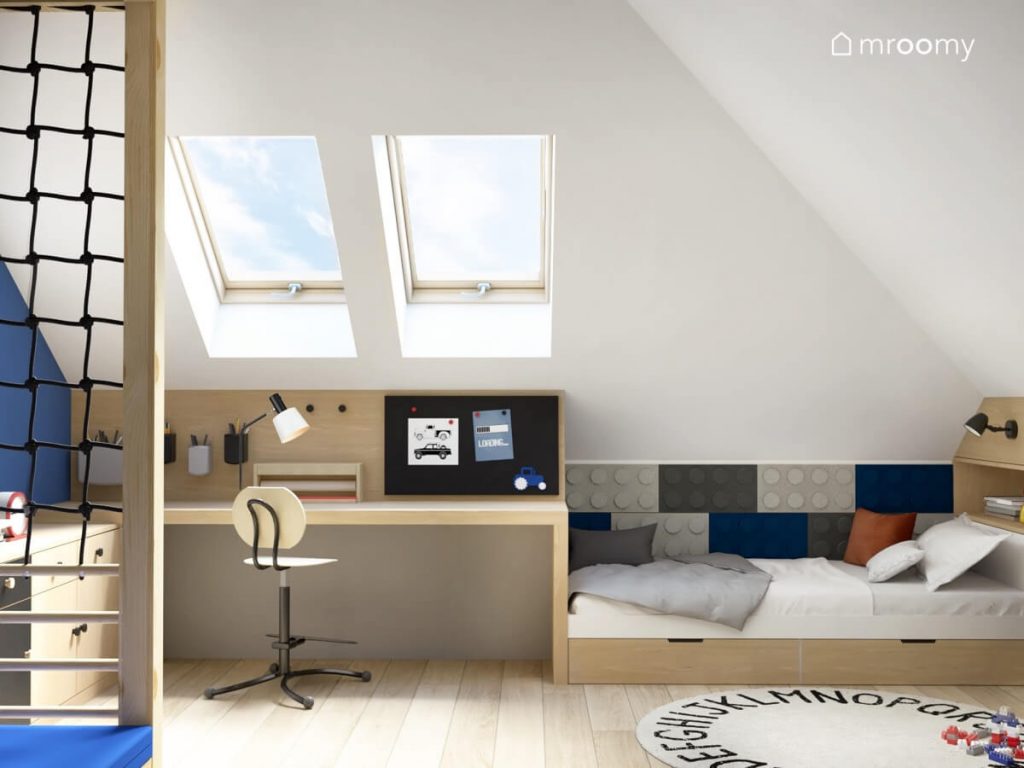 Biało niebieski poddaszowy pokój dla chłopca a w nim drewniane biurko z krzesłem z podnóżkiem łóżko uzupełnione panelami ściennymi w kształcie klocków oraz dywan z alfabetem