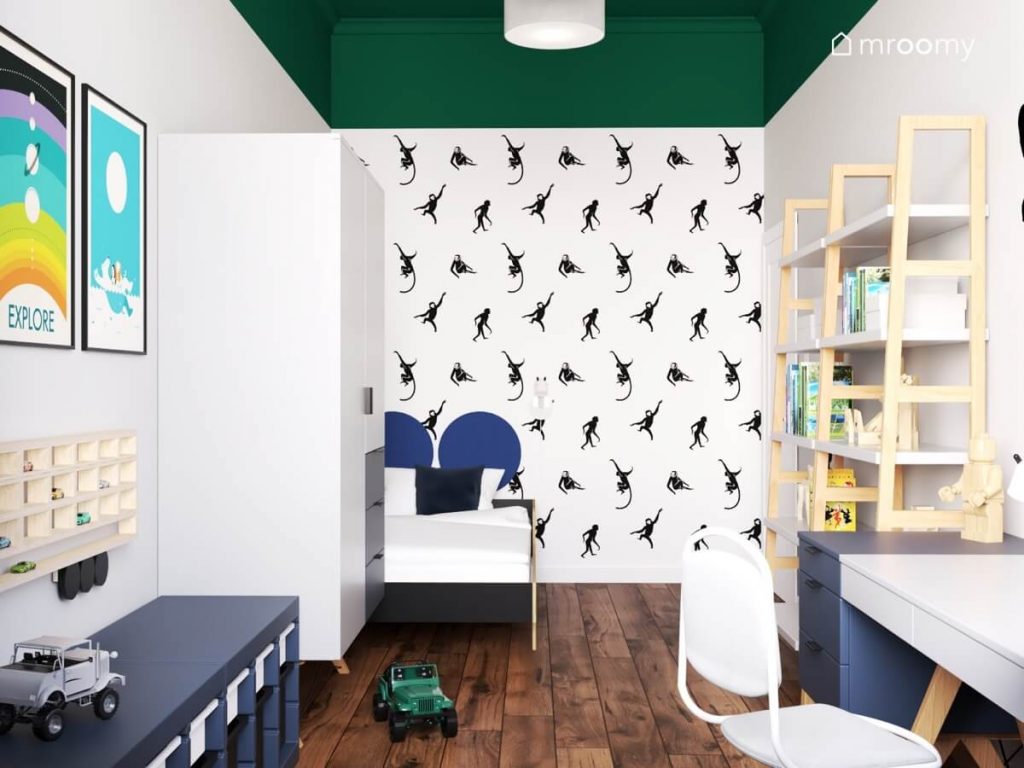 Biały pokój dla chłopca z zielonym sufitem tapetą w małpki na jednej ze ścian oraz biało granatowymi meblami