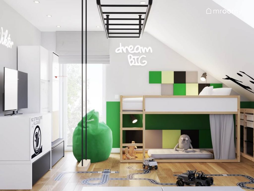 Biało drewniana antresola uzupełniona kwadratowymi panelami w różnych kolorach a obok zielona pufa a na środku prosta huśtawka w pokoju chłopca