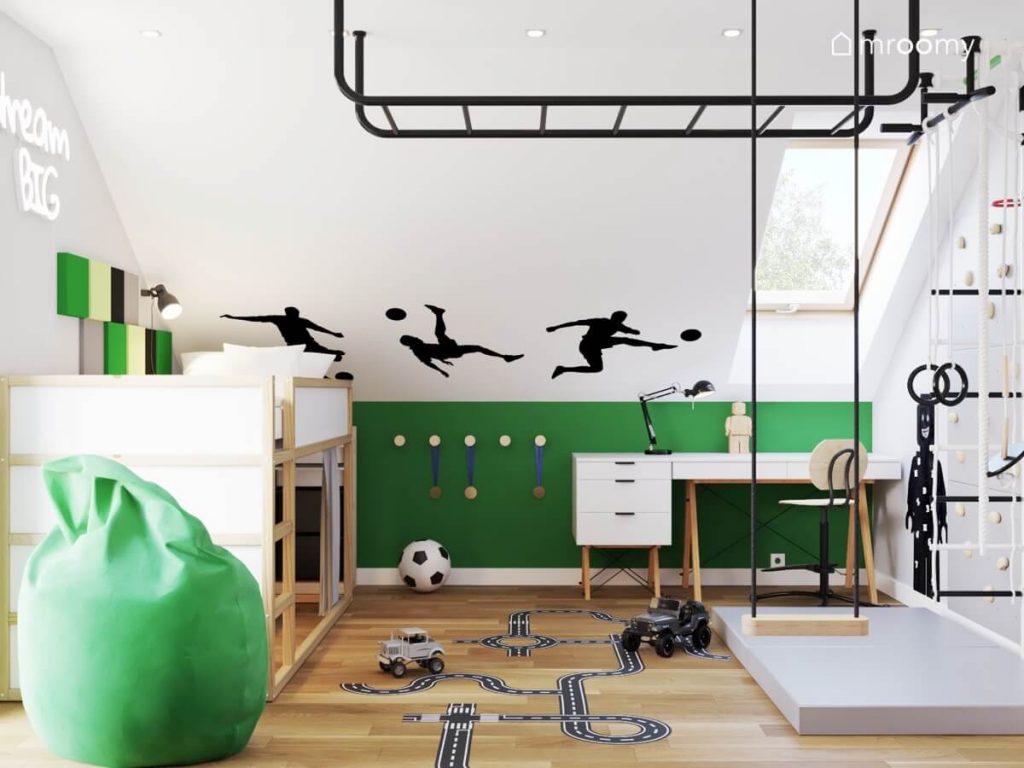 Biało zielony poddaszowy pokój dla chłopca a w nim czarna drabinka sufitowa huśtawka pokojowa naklejki z piłkarzami na skosie oraz miejsce na medale